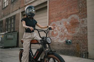 A person wearing a black helmet and casual clothing sits on a dark electric bike in an urban alleyway with a brick wall.